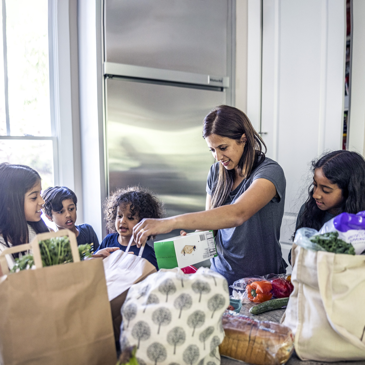 A mother is putting away groceries in the kitchen while her four children observe with curiosity. She has a relaxed expression as she inspects a box of crackers.
